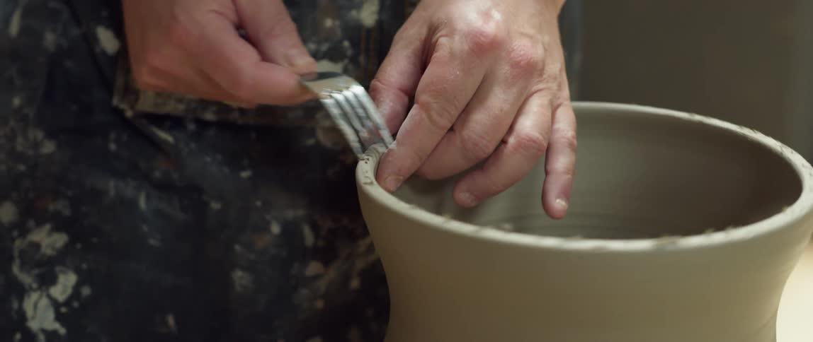 A person uses a fork to score the rim of a piece of pottery as the clays spins on a wheel