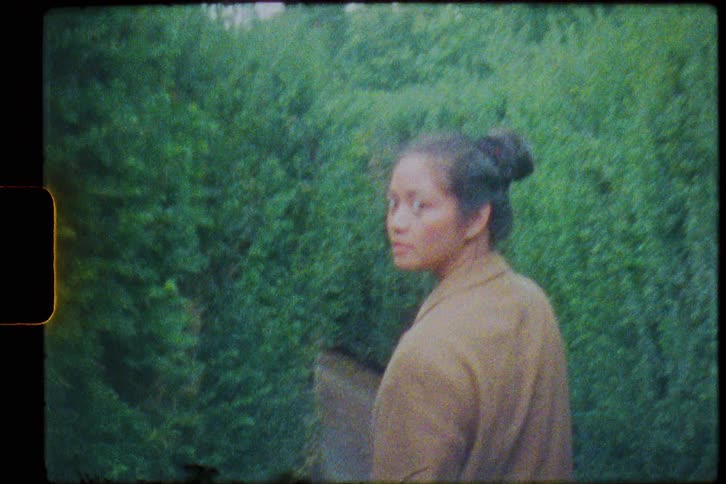 A young woman walking through a hedge maze turns to glance back at the camera as she continues forward