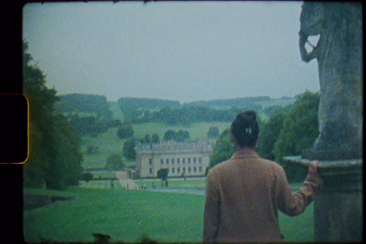 A woman standing with her hand on a statue pedestal looking down at chatsworth house and gardens