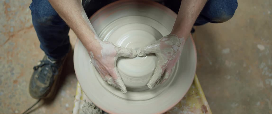 Man sits forming bowl from clay spinning on potters wheel