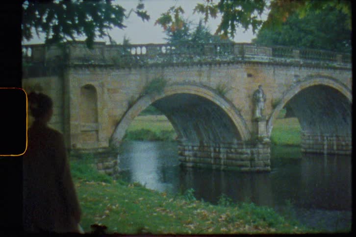 A woman approaches the shore of a river while looking at nearby stone bridge spanning it
