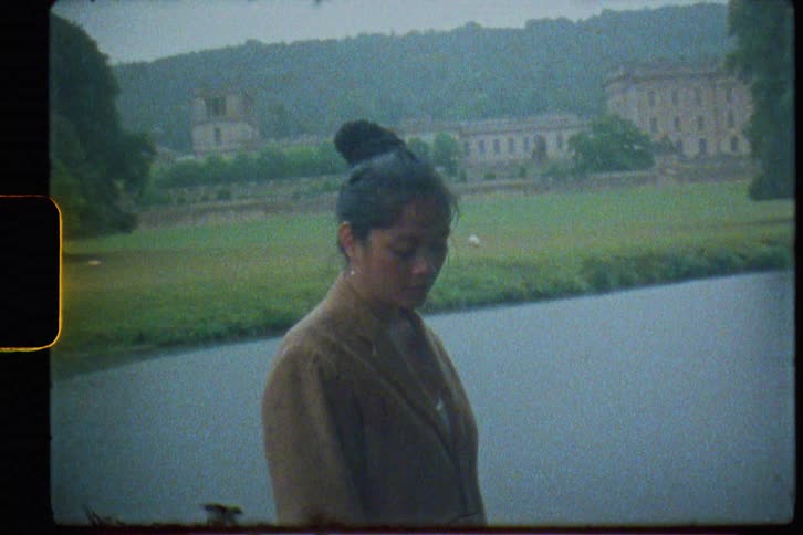 A woman standing on the shore of a river raises her head to look into the camera