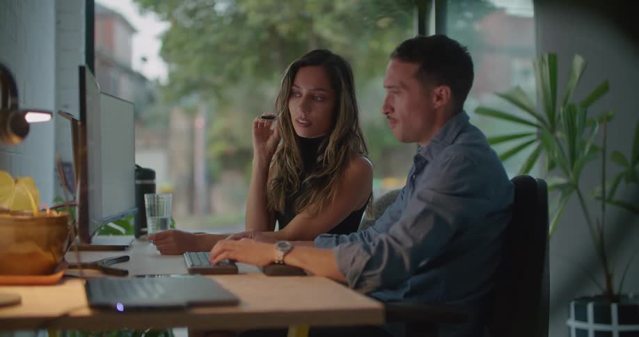 A man and a woman are sitting at a desk in front of a computer and he is pointing to something on the monitor and they talk and she writes something down on paper