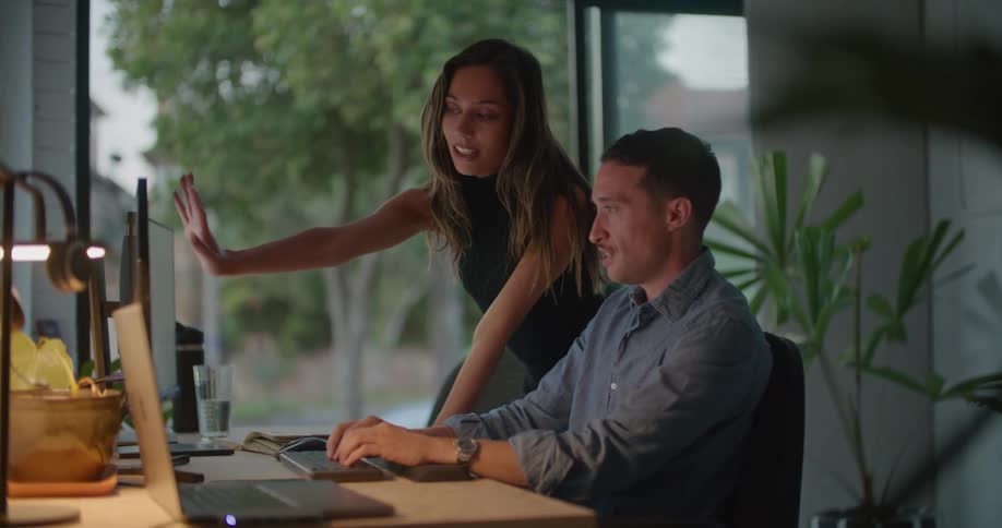 Businessman with mustache and blue shirt sits at desk using computer talking to businesswoman with brown hair and black top in the office