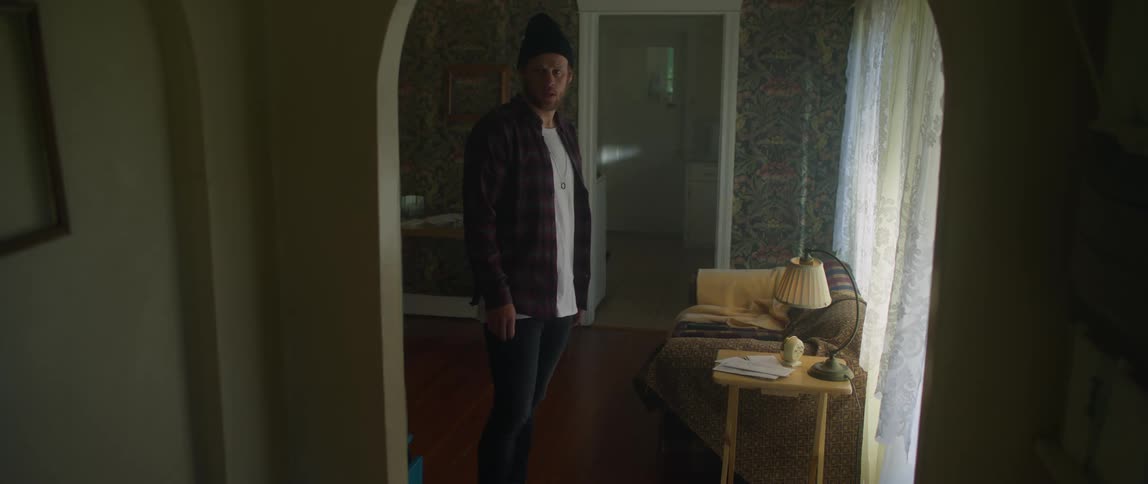 A man wearing a flannel shirt and a knit cap is walking through a room and rushes down a hallway