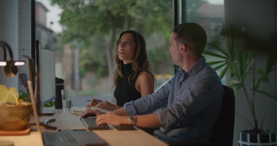 Coworkers sit at desk conversing before one starts typing on computer keyboard