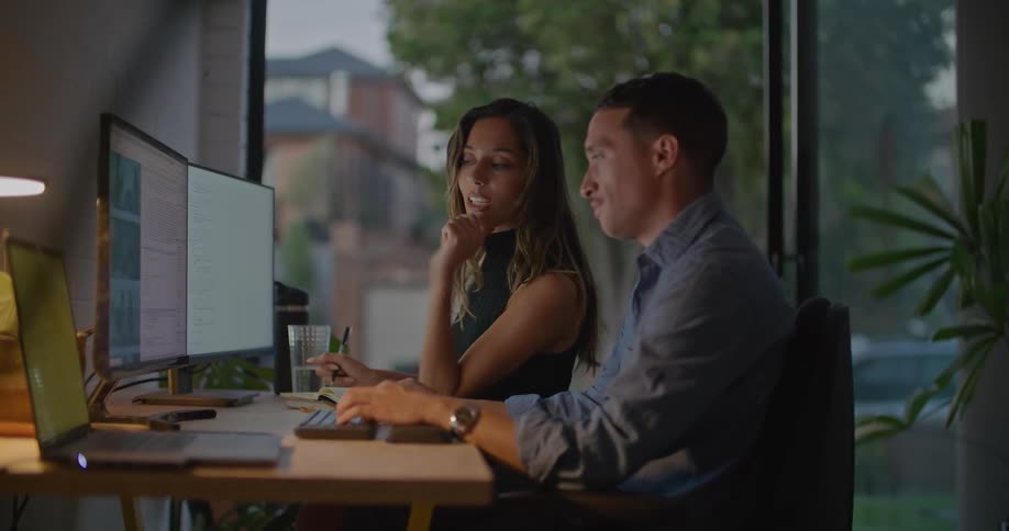 A man is sitting at a table typing on a keyboard and a woman sitting next to him is pointing at something on the monitor