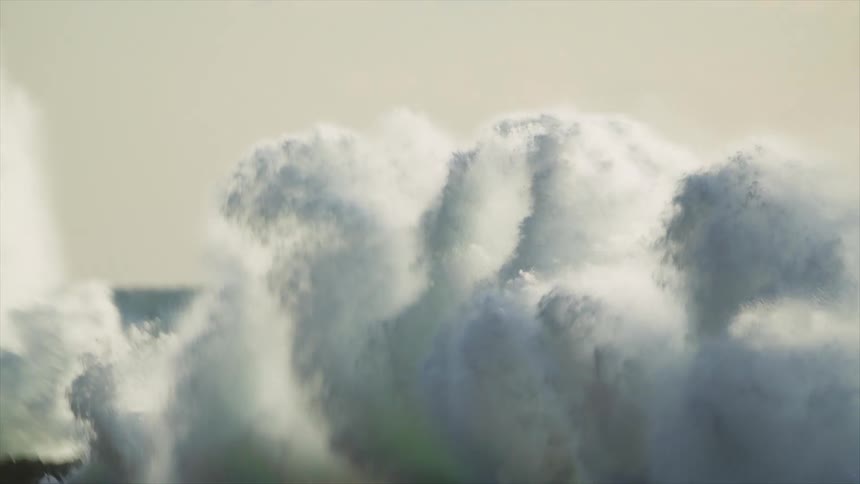 Rough water waves crash against rocks along the shore and splash into the air