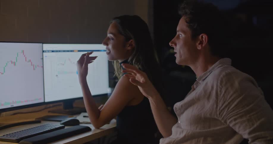 Coworkers sit in office at night discussing graphs showing on computer monitors
