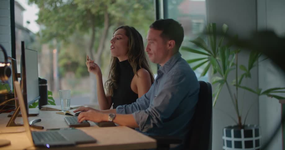 A man and a woman are sitting at a desk in front of a computer and his hands are on the keyboard and she is holding a pen in her hand as she talks