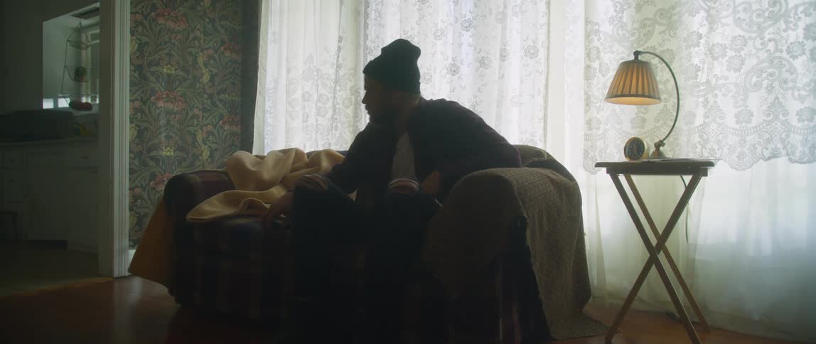A man wearing a flannel shirt and a knit cap is sitting on a living room couch and he picks up a remote controller and sets it back down and gets up and walks away