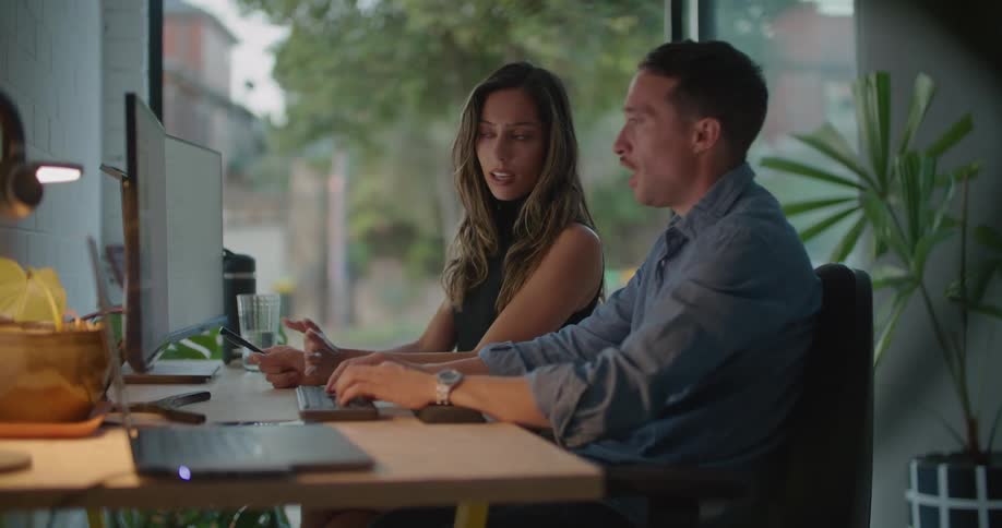 Coworkers sit at desk discussing contents of computer screen near large glass window