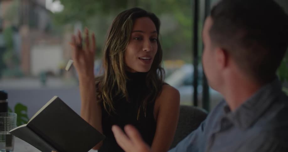 A woman is sitting next to a man and she is holding a book in one hand and a pen in the other as she talks to him