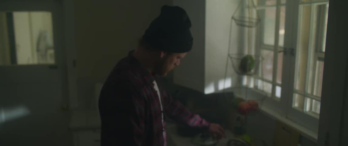 A man wearing a flannel shirt and a knit cap walks into a kitchen and looks around and something startles him