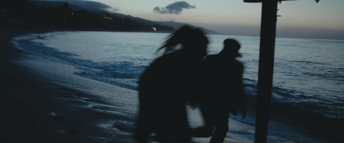 Silhouetted couple move under pier as lights shine in dark town in the background