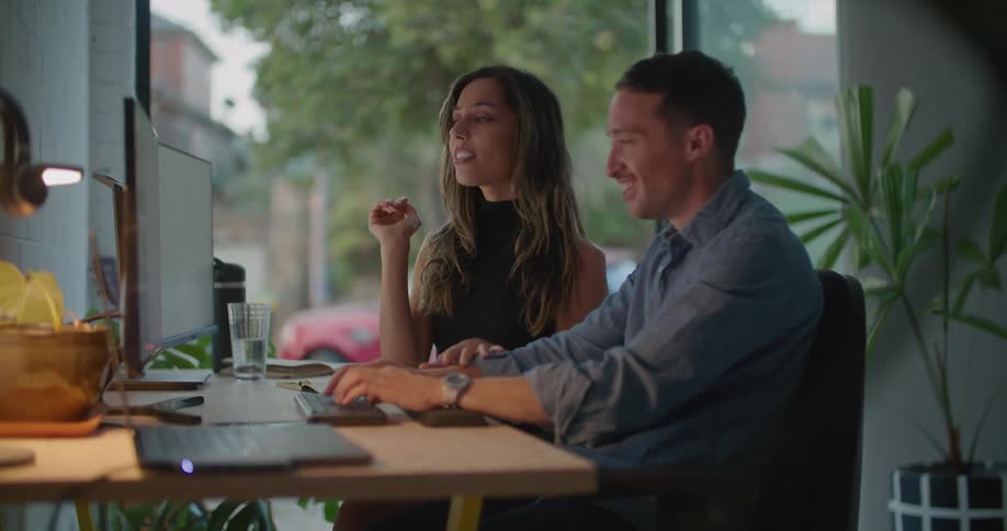 A man typing on a computer talks with the woman sitting next to him at the computer desk