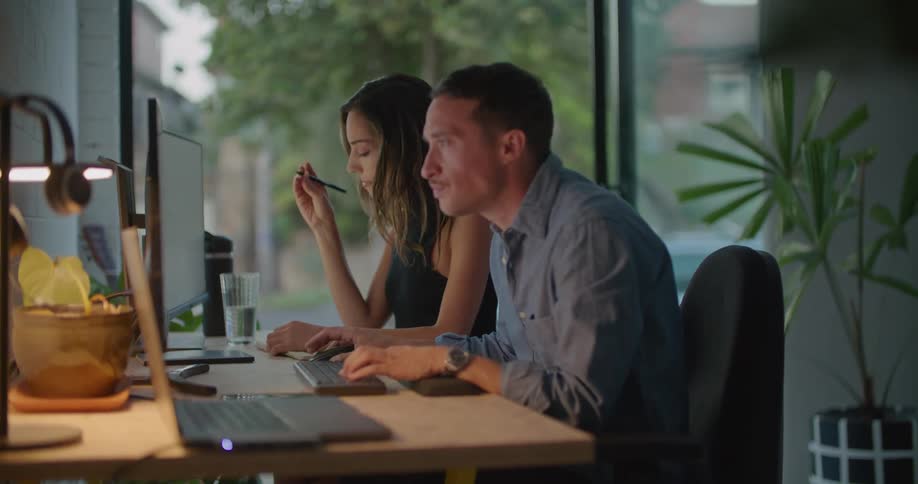 A man is sitting at a table typing on a keyboard and a woman sitting next to him points at something on the monitor