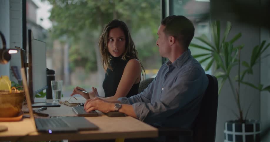 A man typing on a computer talks with a woman writing in a notebook next to him