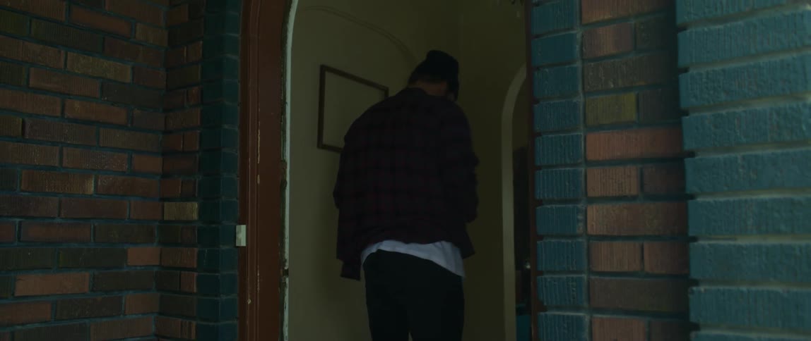 A man wearing a flannel shirt and a knit cap bursts out of the front door of a house looking around and picks up a letter from the ground and runs back out after reading it