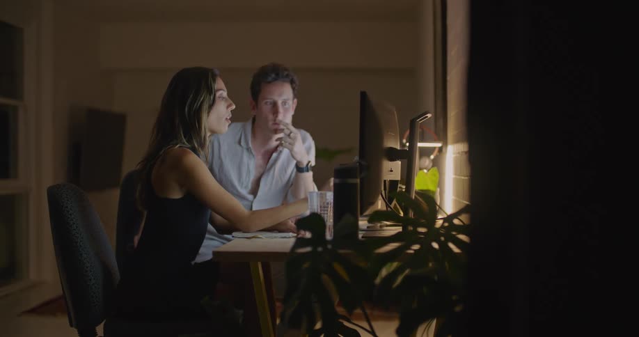 A woman and man sitting before a computer at a desk gesture at the monitor while talking to one another