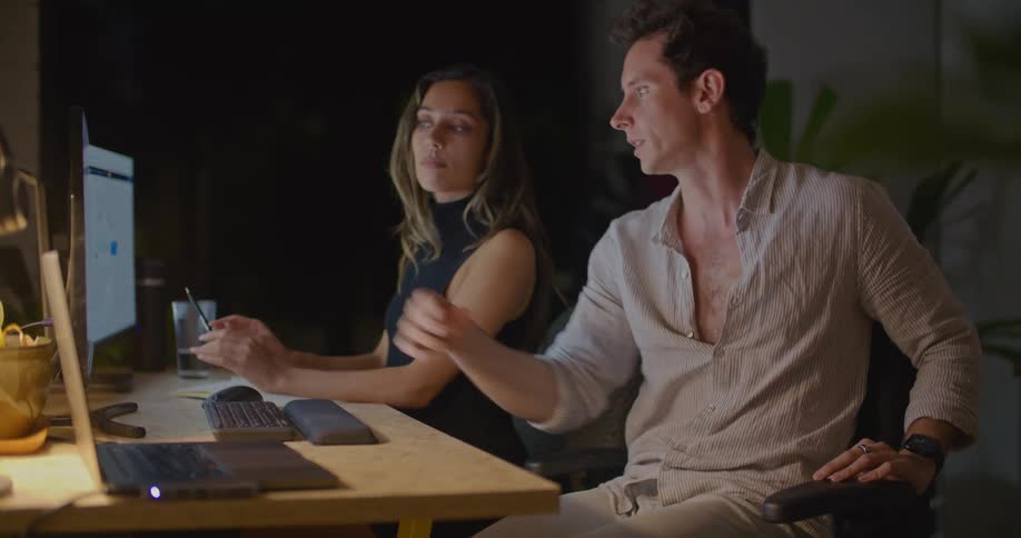A man and woman sitting before a computer at a desk talk with one another while gesturing at the monitors