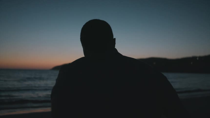 Silhouette of a man standing on a beach taking off his jacket as the waves roll over the sand
