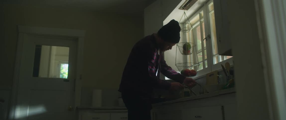 A man stands in his kitchen trying to turn the water at the sink on and then walks in to another room and picks up a chair that has been knocked down as he looks around