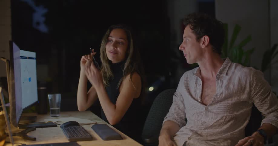 Coworkers sits at desk near sliding glass doors at night discussing data on screen