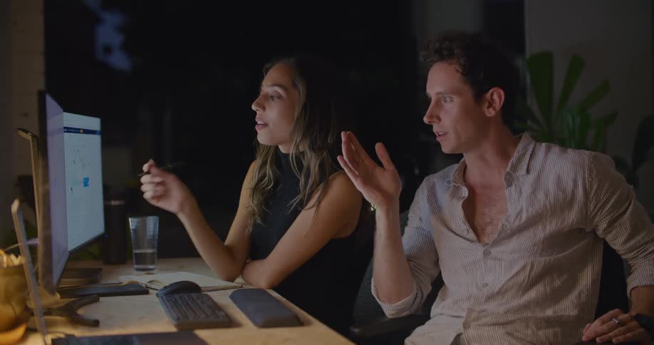 A man and woman sit at a desk in an office and brainstorm as they look at computer monitors