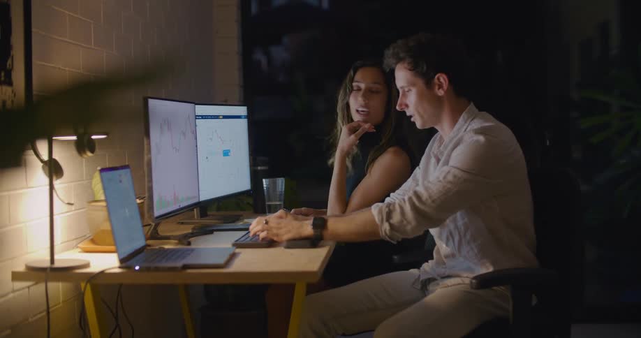Two coworkers sit at a desk and talk as they discuss information on computer screens