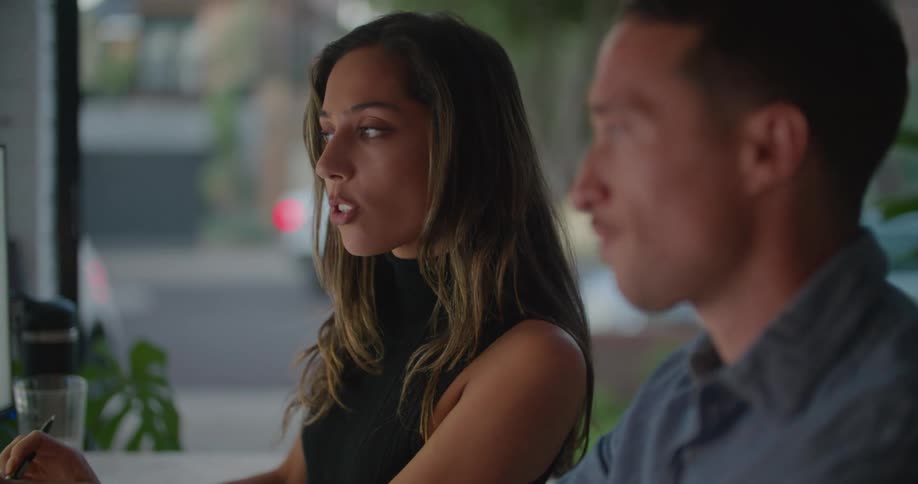 A man and woman sit at a desk together and the man points to a computer screen and they talk
