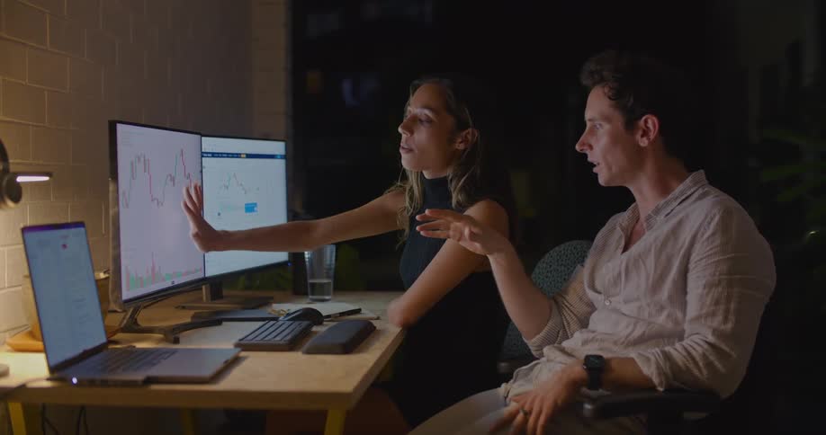 A man and a woman are sitting at a desk in front of computer monitors and she is pointing to the monitors explaining something and he sits back in his chair and talks to her