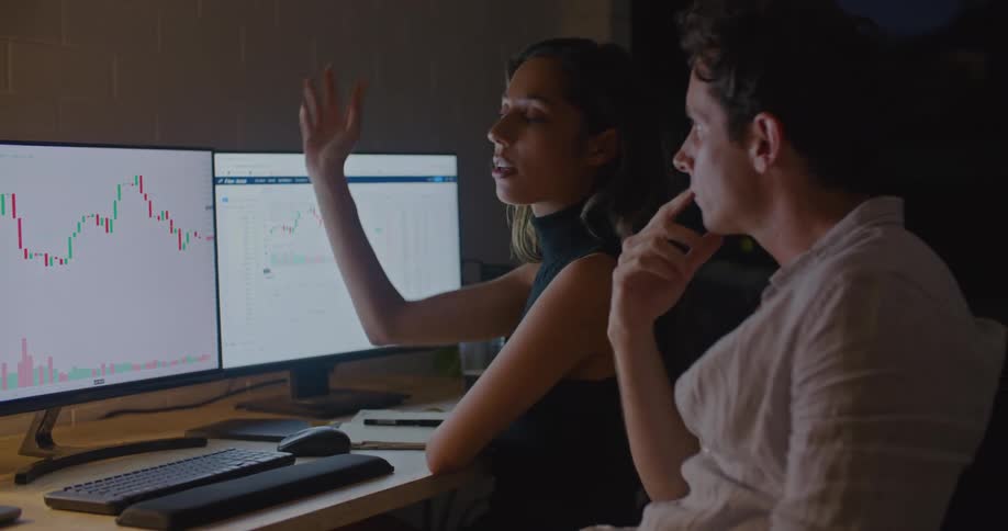 Coworkers sit at desk together at night discussing data graphed on computer display
