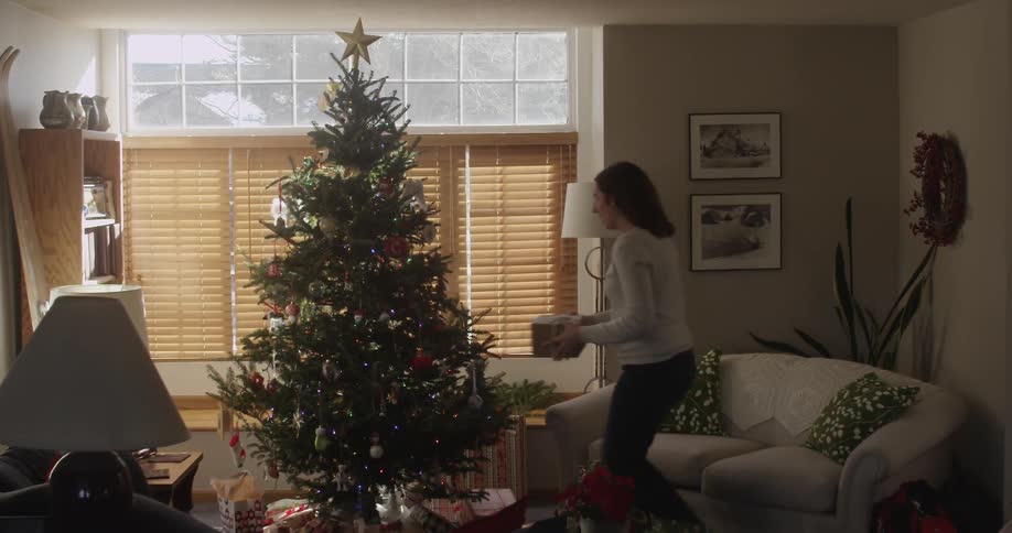 A woman walks over to a christmas tree and places a gift underneath
