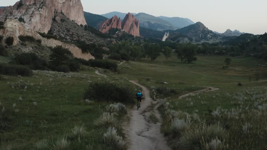 A cyclist rides a bicycle on a trail near rock formations and there are mountains in the distance