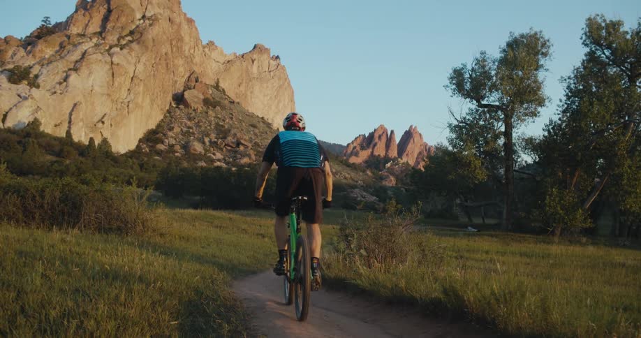A man rides a bike on a curving narrow trail towards jagged rock formations