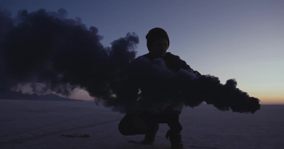 A man squatting holding a smoke flare at dusk