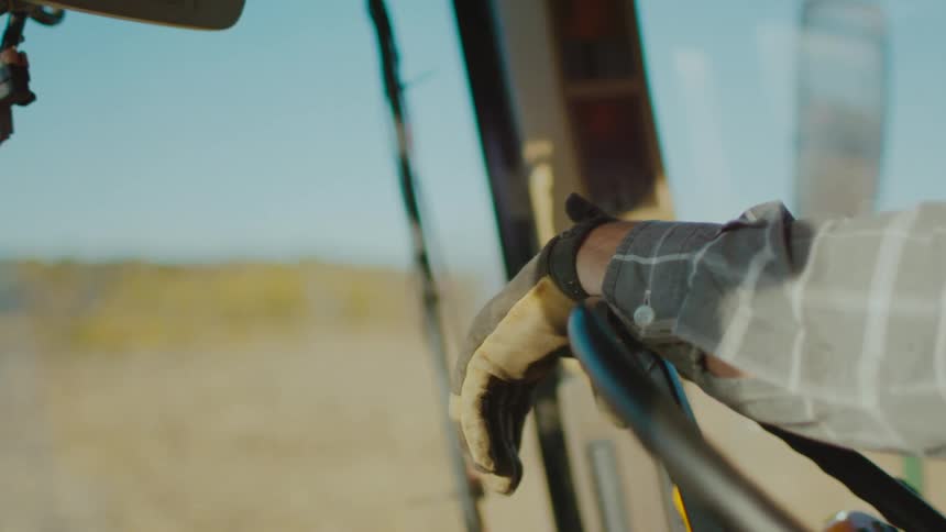 Slow motion shot of a gloved hand and sleeved arm resting on the steering wheel of a large farm vehicle as it moves along a flat plain overlooking nothing but yellow fields extending all the way to the horizon under a cloudless pale blue sky