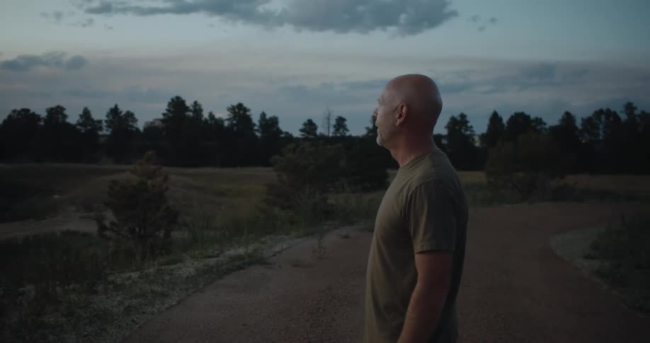 Bald middle aged man with green t shirt stands on dirt road looking around at dusk