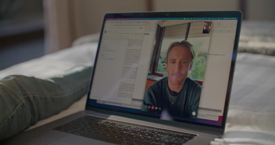 A person in a room sitting on a bed looking at a laptop with a wall in the background