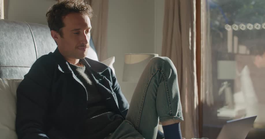 A man is sitting on a bed and talking to someone on a laptop