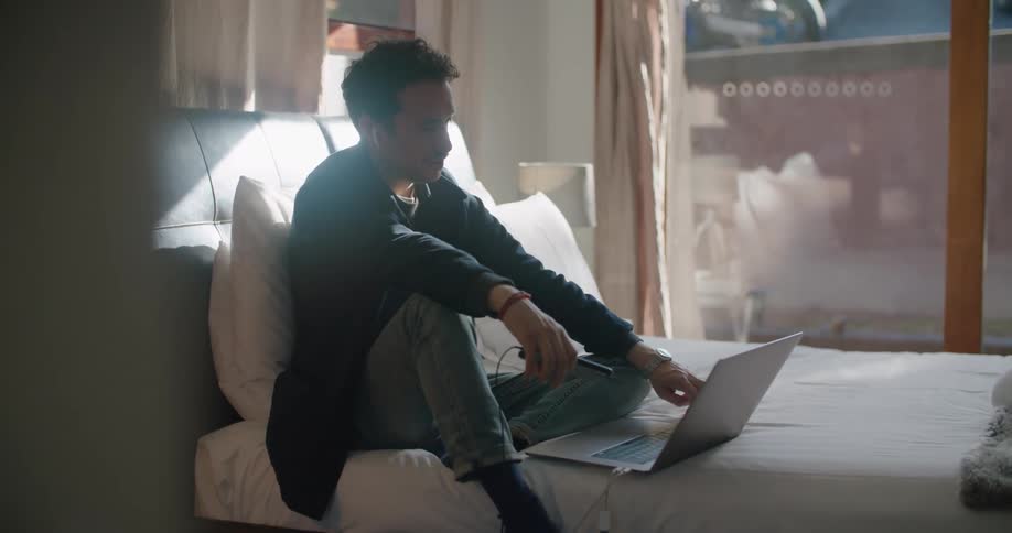 Man sits on a bed with a laptop and holds a cellphone as a motorcycle sits outside