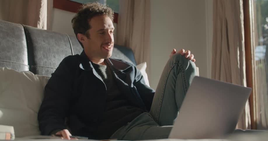 A man uses a laptop to participate in a video chat while sitting on a bed