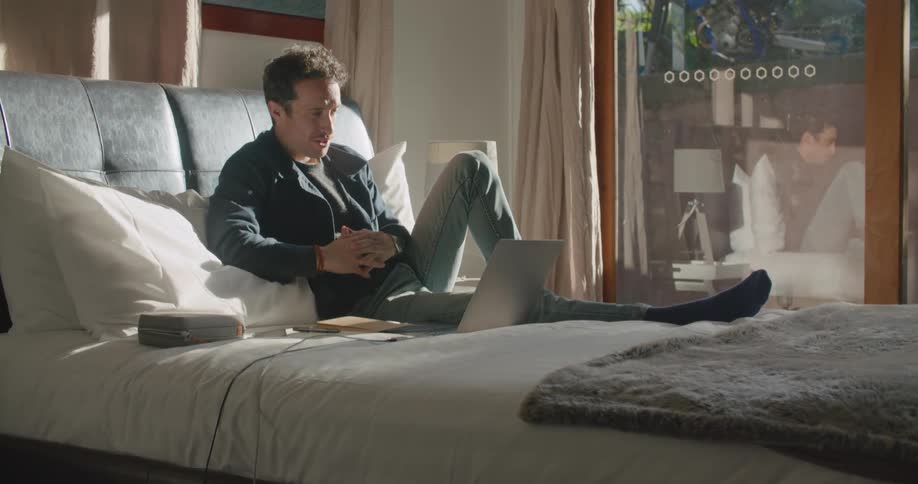 Man with dark jacket and jeans sits on bed making a video call on laptop in the bedroom