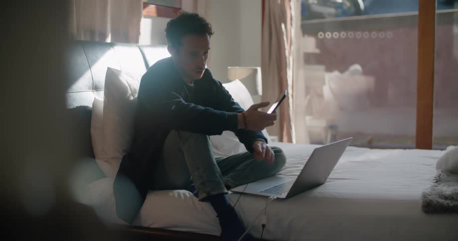 A man talks on his smartphone while sitting in bed and using his laptop