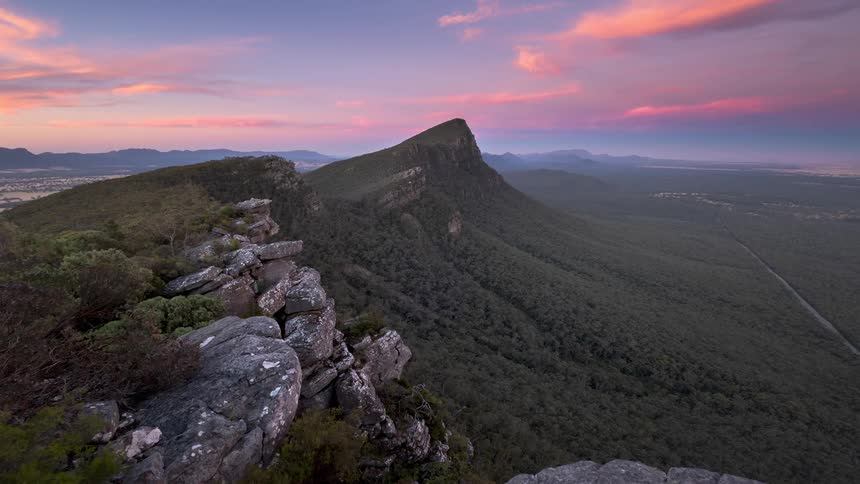 Rocks along a mountain with peaks high in the sky as clouds pass by and there are mountains on the horizon