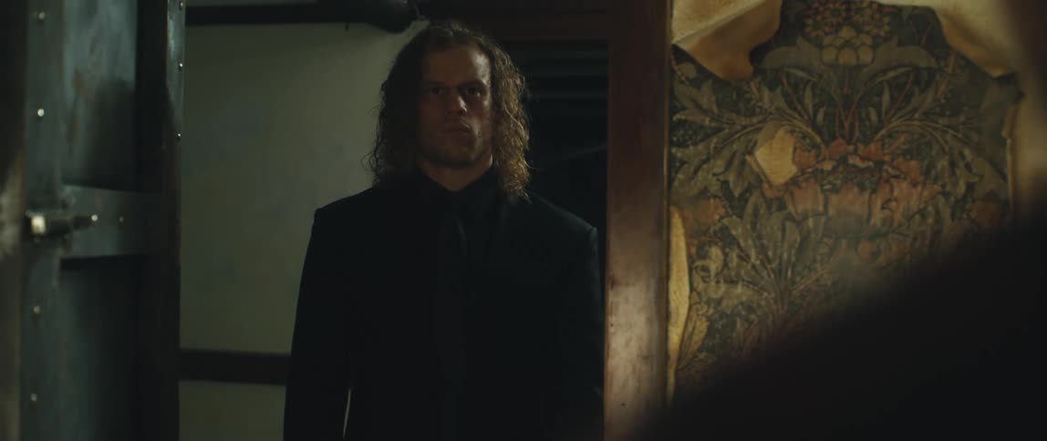 Man with long curly hair and black suit stares at man and shutting the door on man