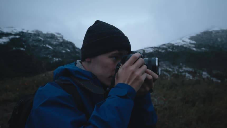A photographer raises his camera to take a photograph while standing in the mountains on a cloudy day