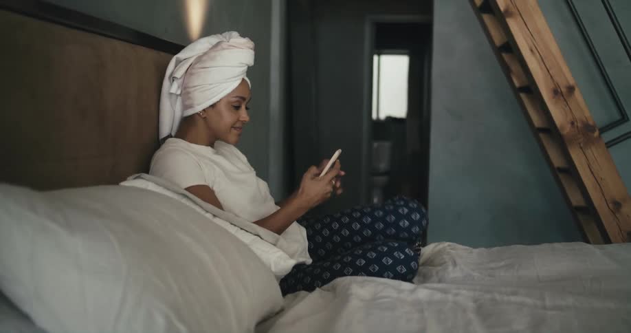 A woman with a towel around her head sits on a bed and uses her phone