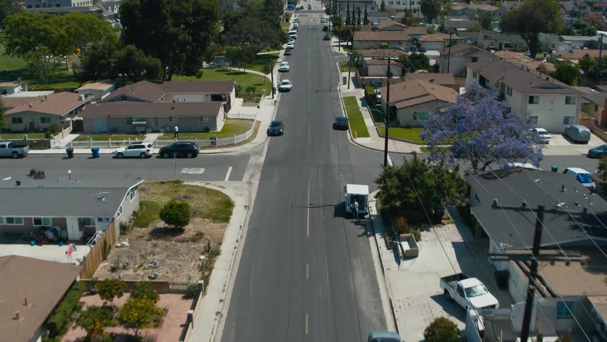 An aerial view of a neighborhood with cars parked on the streets and a man walking across a lawn towards a house